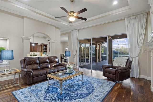 living room with dark wood-type flooring, a raised ceiling, crown molding, ceiling fan, and ornate columns