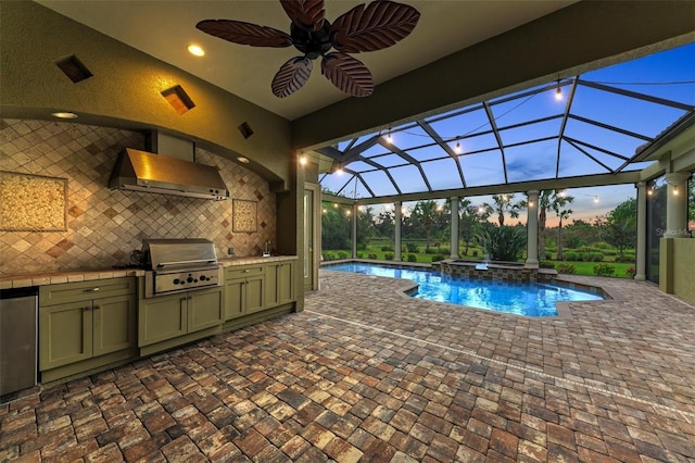 pool at dusk featuring glass enclosure, grilling area, ceiling fan, a patio area, and exterior kitchen