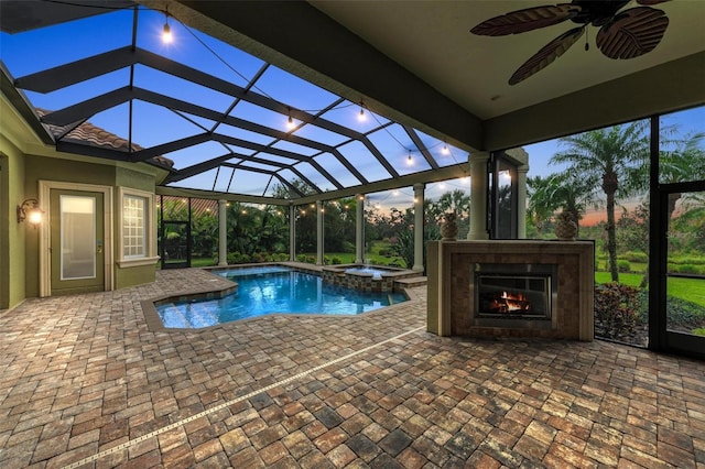 pool at dusk featuring glass enclosure, ceiling fan, exterior fireplace, an in ground hot tub, and a patio