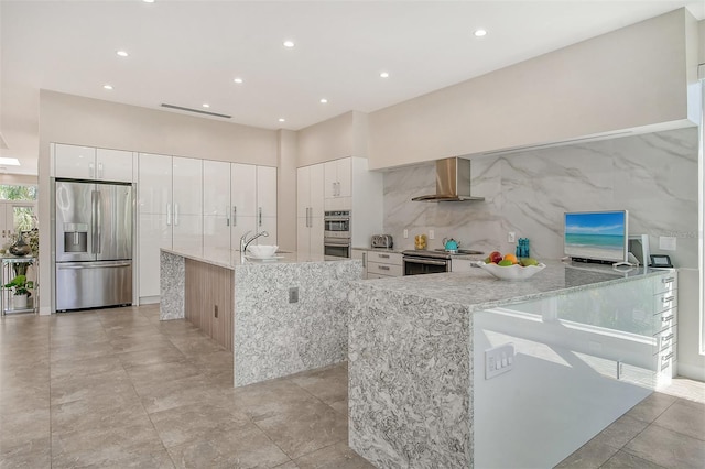 kitchen with appliances with stainless steel finishes, light stone counters, wall chimney exhaust hood, sink, and white cabinetry