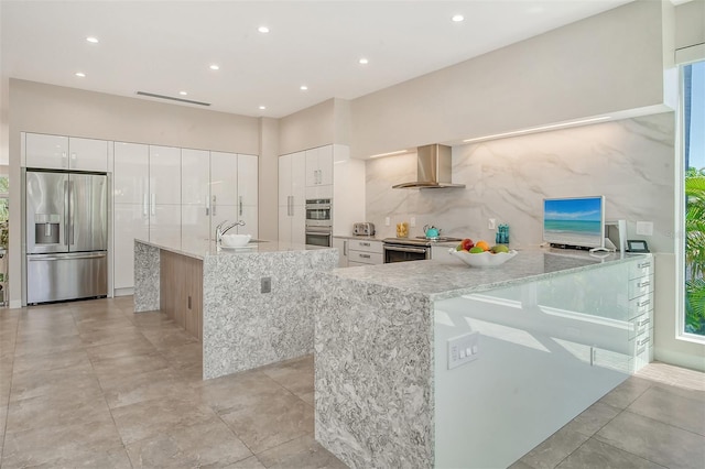 kitchen with wall chimney exhaust hood, light stone counters, white cabinetry, and appliances with stainless steel finishes