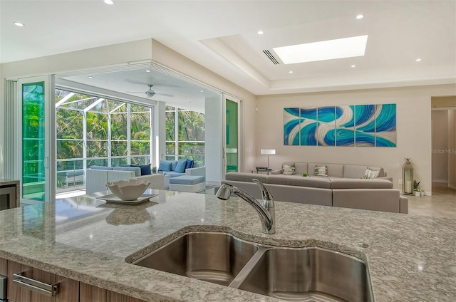 kitchen with light stone countertops, a skylight, a tray ceiling, ceiling fan, and sink