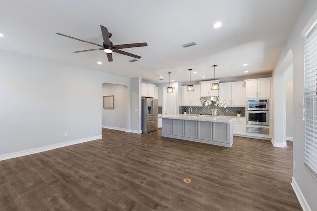 kitchen featuring arched walkways, stainless steel appliances, white cabinets, a center island with sink, and pendant lighting