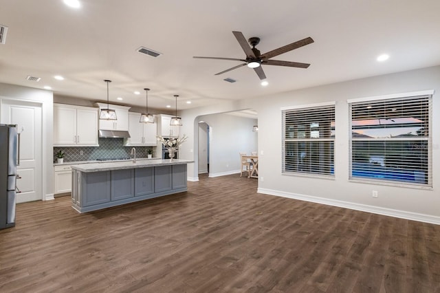 kitchen featuring arched walkways, an island with sink, hanging light fixtures, under cabinet range hood, and white cabinetry