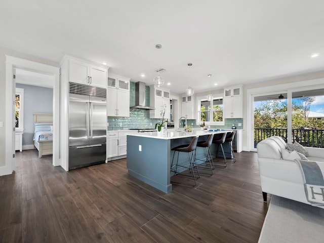 kitchen featuring wall chimney range hood, dark hardwood / wood-style floors, built in refrigerator, decorative light fixtures, and a center island with sink