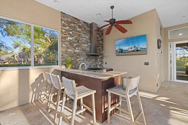 bar featuring ceiling fan, wall chimney exhaust hood, stone tile floors, and a textured ceiling