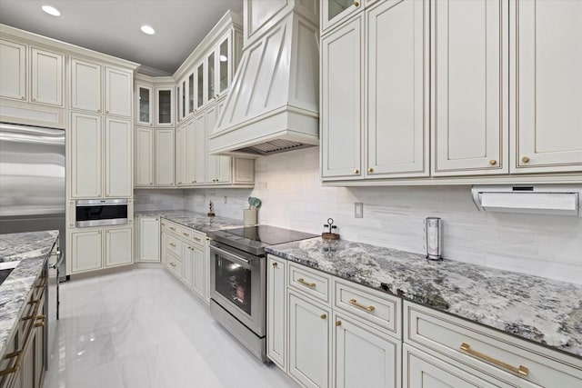 kitchen with tasteful backsplash, appliances with stainless steel finishes, glass insert cabinets, light stone counters, and custom exhaust hood