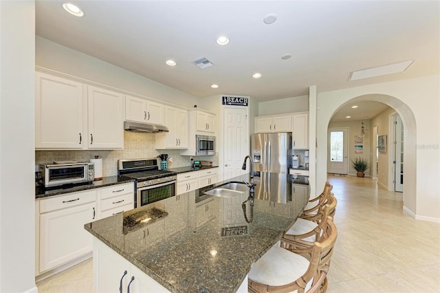 kitchen with sink, white cabinets, dark stone counters, stainless steel appliances, and a center island with sink