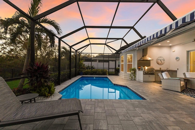 pool at dusk featuring area for grilling, a patio area, exterior kitchen, and glass enclosure