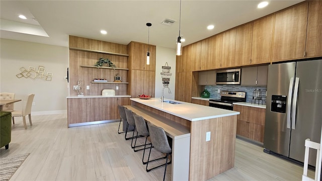 kitchen with tasteful backsplash, stainless steel appliances, decorative light fixtures, a center island with sink, and light hardwood / wood-style floors