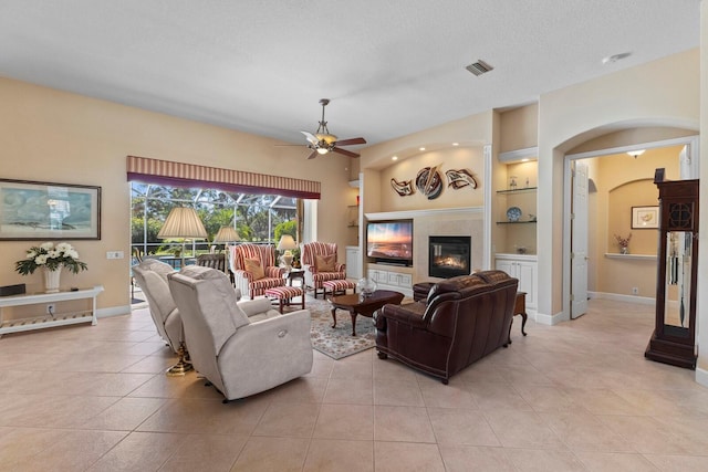 living room featuring a tile fireplace, ceiling fan, built in features, a textured ceiling, and light tile patterned flooring