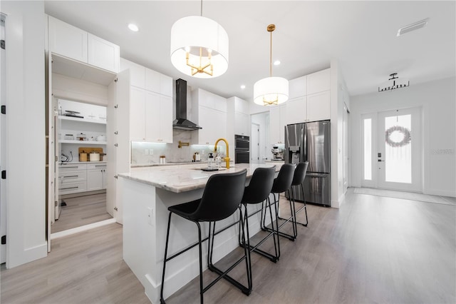 kitchen with stainless steel fridge, wall chimney range hood, a center island with sink, white cabinets, and hanging light fixtures