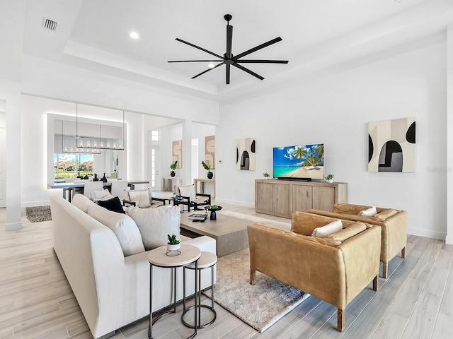 living room featuring ceiling fan with notable chandelier, a towering ceiling, light wood-type flooring, and a raised ceiling