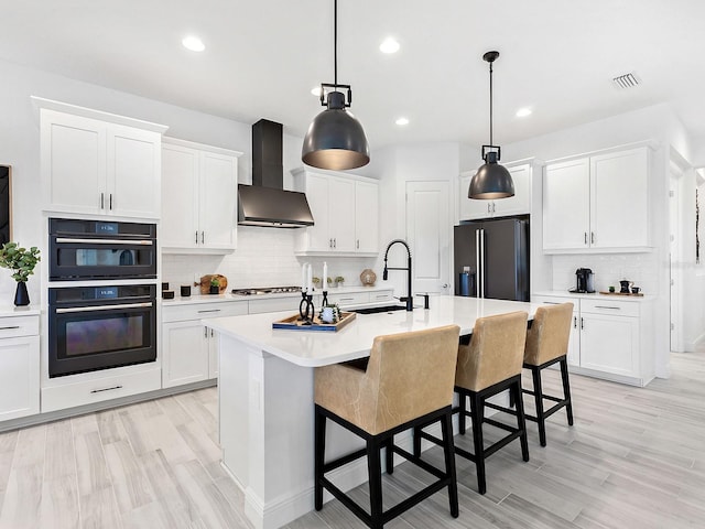 kitchen with hanging light fixtures, stainless steel appliances, wall chimney range hood, an island with sink, and decorative backsplash