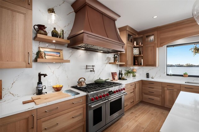 kitchen with light stone countertops, premium range hood, light hardwood / wood-style floors, range with two ovens, and decorative backsplash