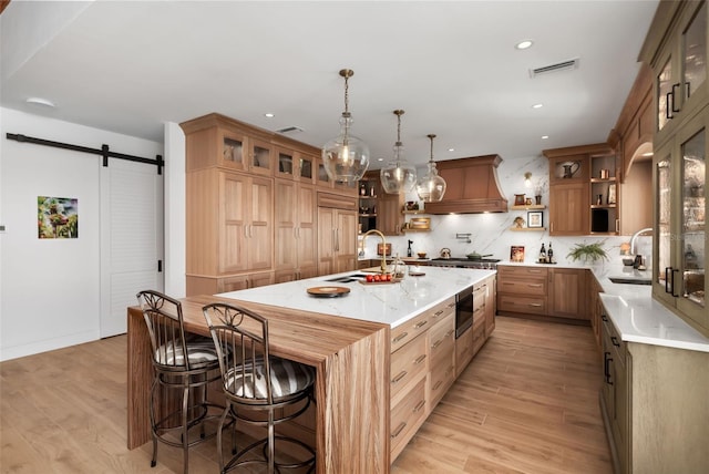 kitchen featuring premium range hood, sink, a barn door, light wood-type flooring, and an island with sink