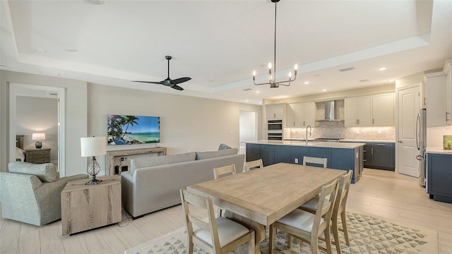 dining area featuring ceiling fan with notable chandelier, a raised ceiling, light wood-type flooring, and sink