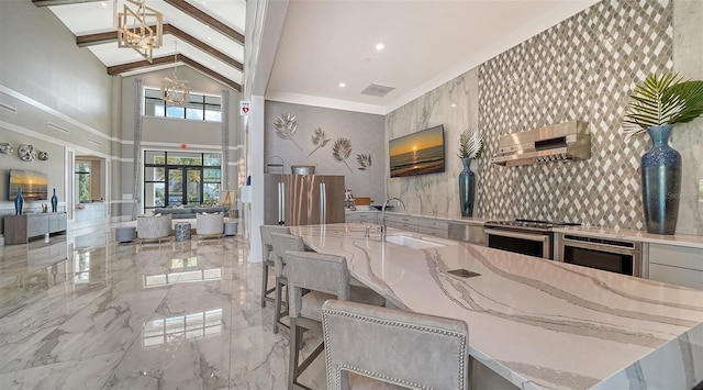 kitchen with beam ceiling, stainless steel appliances, wall chimney range hood, light stone counters, and a notable chandelier