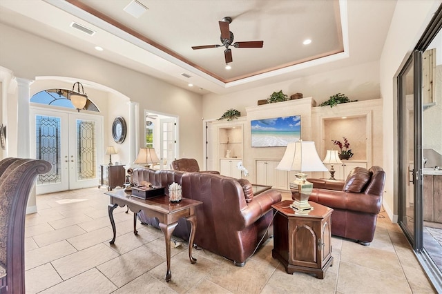 tiled living room with ceiling fan, ornate columns, a tray ceiling, and french doors