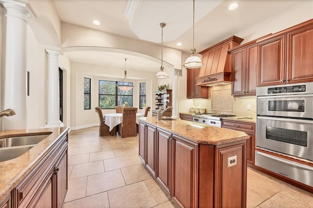 kitchen with sink, stainless steel appliances, hanging light fixtures, decorative columns, and an island with sink