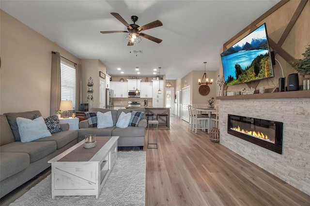 living room with light wood-type flooring, ceiling fan with notable chandelier, and a stone fireplace