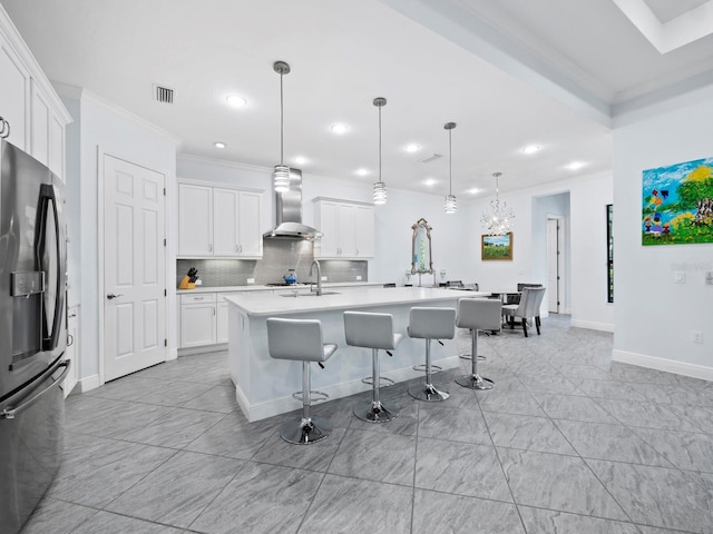 kitchen featuring hanging light fixtures, stainless steel fridge with ice dispenser, wall chimney exhaust hood, an island with sink, and white cabinetry