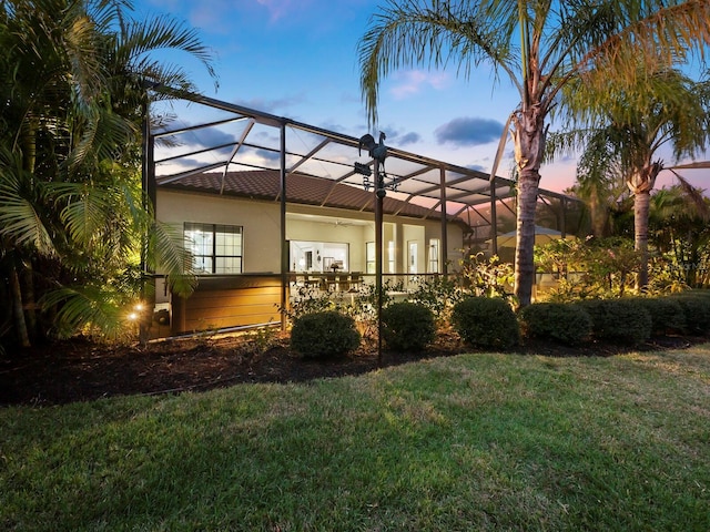 back house at dusk featuring glass enclosure, ceiling fan, and a lawn