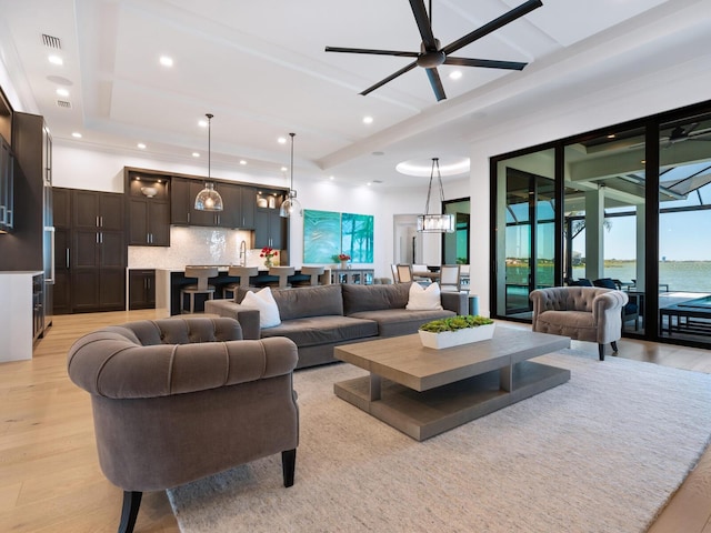 living room featuring a raised ceiling, ceiling fan with notable chandelier, light hardwood / wood-style floors, and a water view