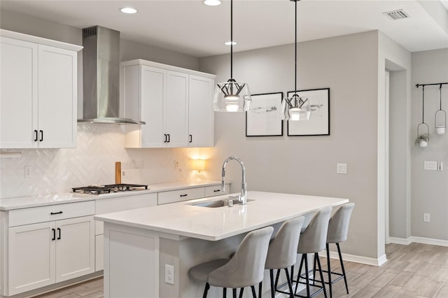 kitchen featuring wall chimney exhaust hood, sink, decorative light fixtures, white cabinets, and an island with sink