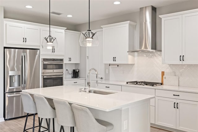 kitchen featuring a center island with sink, wall chimney range hood, sink, hanging light fixtures, and stainless steel appliances