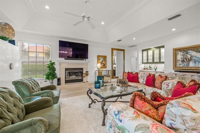 living room with light wood-type flooring, ornamental molding, a tray ceiling, ceiling fan, and a tiled fireplace