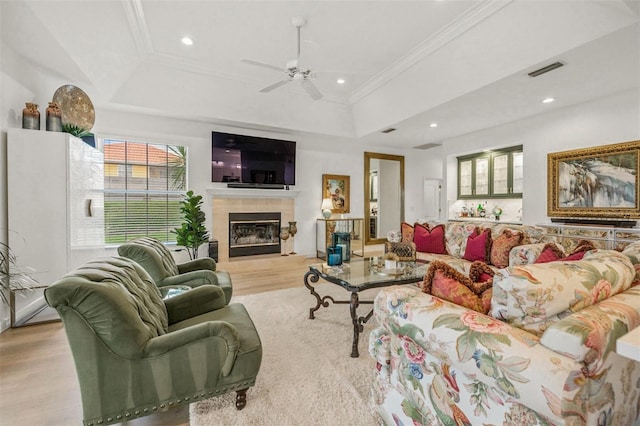 living room featuring a raised ceiling, ceiling fan, crown molding, light hardwood / wood-style flooring, and a tiled fireplace