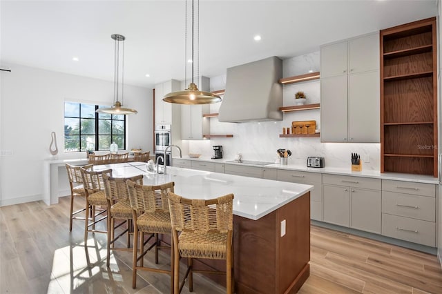 kitchen with exhaust hood, a center island with sink, black electric cooktop, light hardwood / wood-style floors, and a breakfast bar area