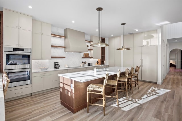 kitchen with pendant lighting, wall chimney range hood, light hardwood / wood-style flooring, an island with sink, and double oven