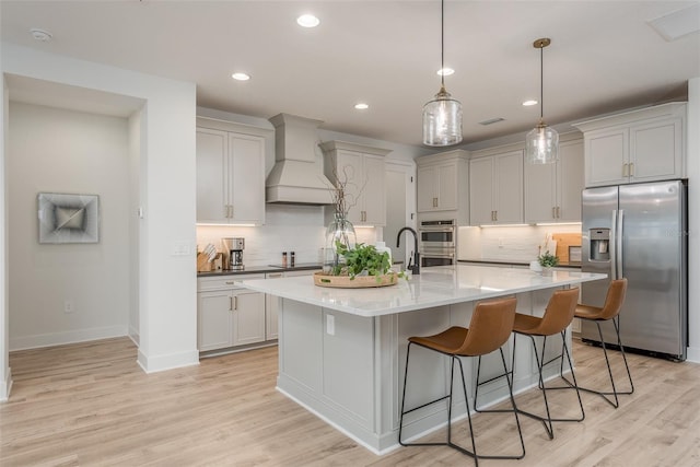 kitchen featuring premium range hood, appliances with stainless steel finishes, a center island with sink, and light hardwood / wood-style flooring