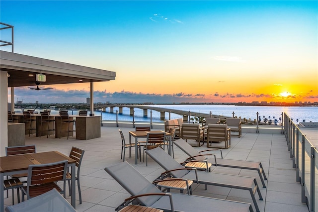 patio terrace at dusk featuring a water view, an outdoor bar, and ceiling fan