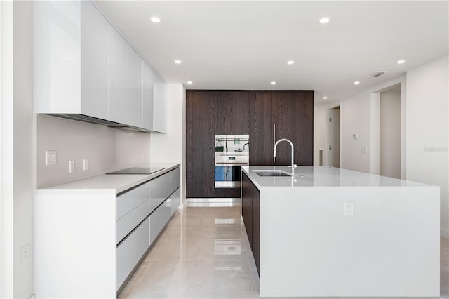 kitchen with an island with sink, sink, stainless steel oven, dark brown cabinets, and black electric cooktop