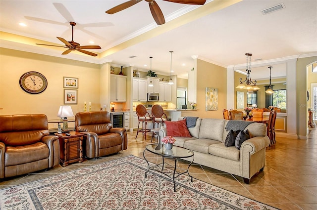 tiled living room with ceiling fan with notable chandelier, wine cooler, and ornamental molding