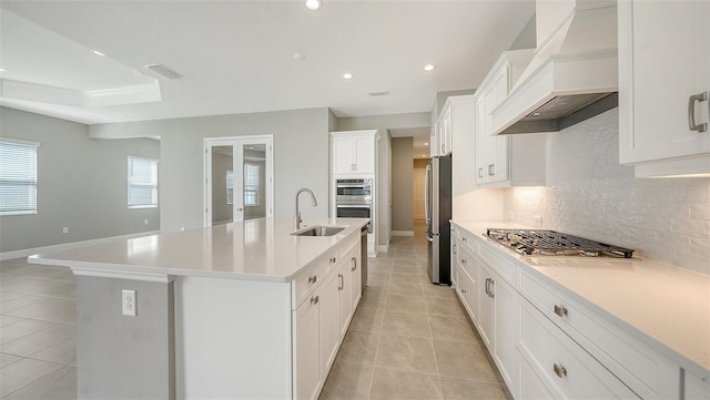 kitchen with appliances with stainless steel finishes, backsplash, premium range hood, a center island with sink, and white cabinetry