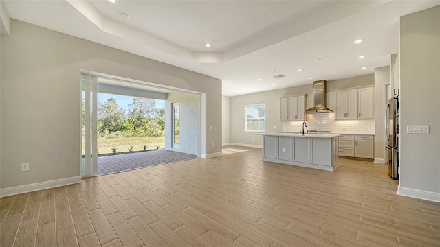 kitchen featuring a center island with sink, wall chimney range hood, light hardwood / wood-style flooring, decorative backsplash, and stainless steel fridge