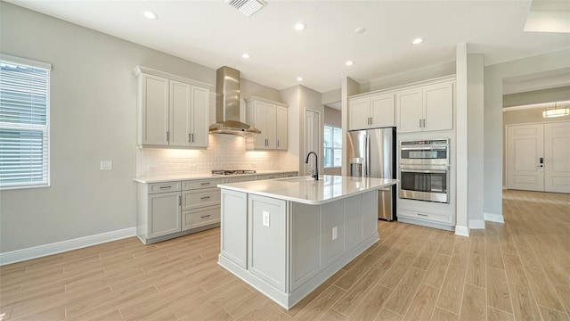 kitchen with light wood-type flooring, stainless steel appliances, wall chimney range hood, white cabinetry, and an island with sink