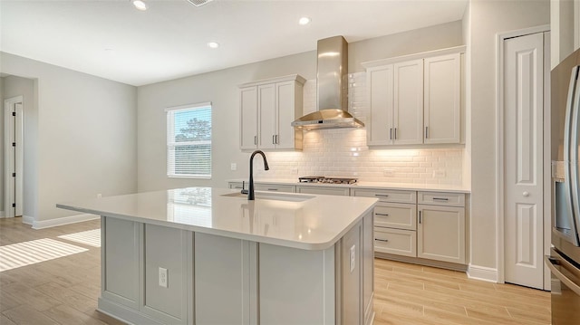 kitchen featuring wall chimney exhaust hood, sink, light wood-type flooring, and an island with sink