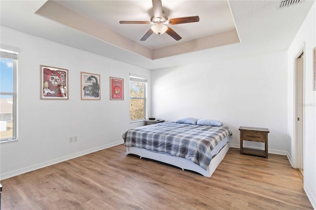 bedroom featuring a raised ceiling, multiple windows, ceiling fan, and light hardwood / wood-style flooring