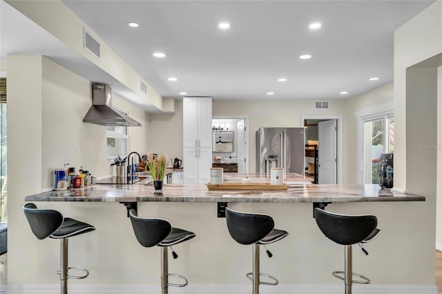 kitchen with kitchen peninsula, stainless steel fridge, wall chimney exhaust hood, a breakfast bar, and white cabinetry