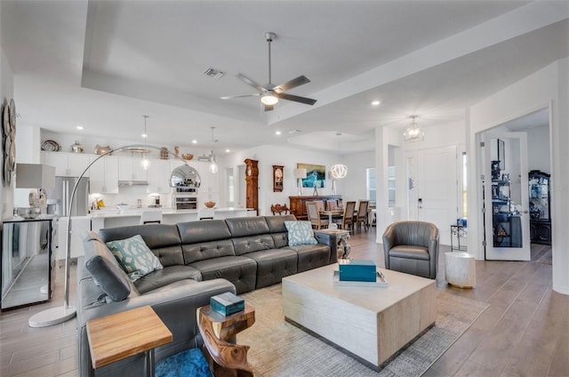 living room featuring ceiling fan with notable chandelier, a tray ceiling, and light hardwood / wood-style flooring