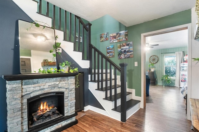 staircase featuring wood-type flooring, a textured ceiling, a stone fireplace, and ceiling fan