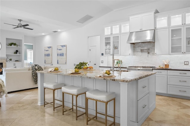 kitchen featuring light stone countertops, vaulted ceiling, exhaust hood, a center island with sink, and white cabinets