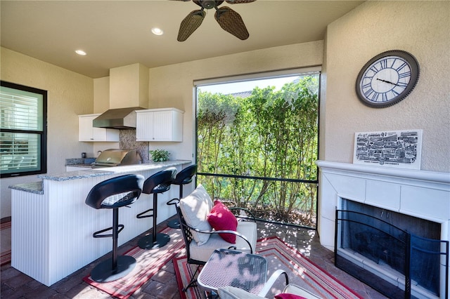 kitchen with a breakfast bar, backsplash, wall chimney range hood, ceiling fan, and white cabinetry