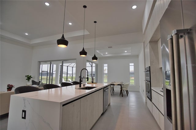 kitchen with light stone countertops, sink, pendant lighting, a tray ceiling, and a center island with sink