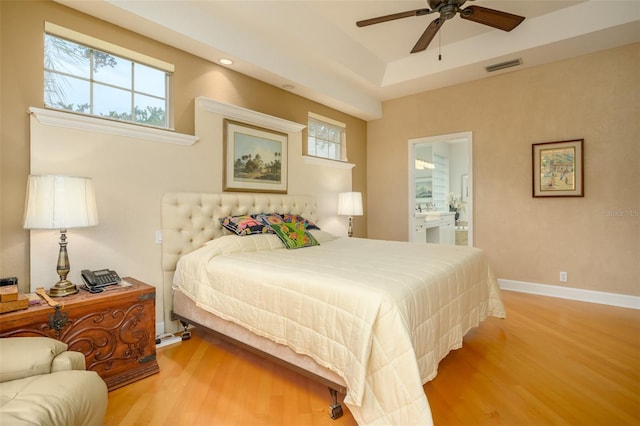 bedroom with a raised ceiling, connected bathroom, ceiling fan, and light wood-type flooring